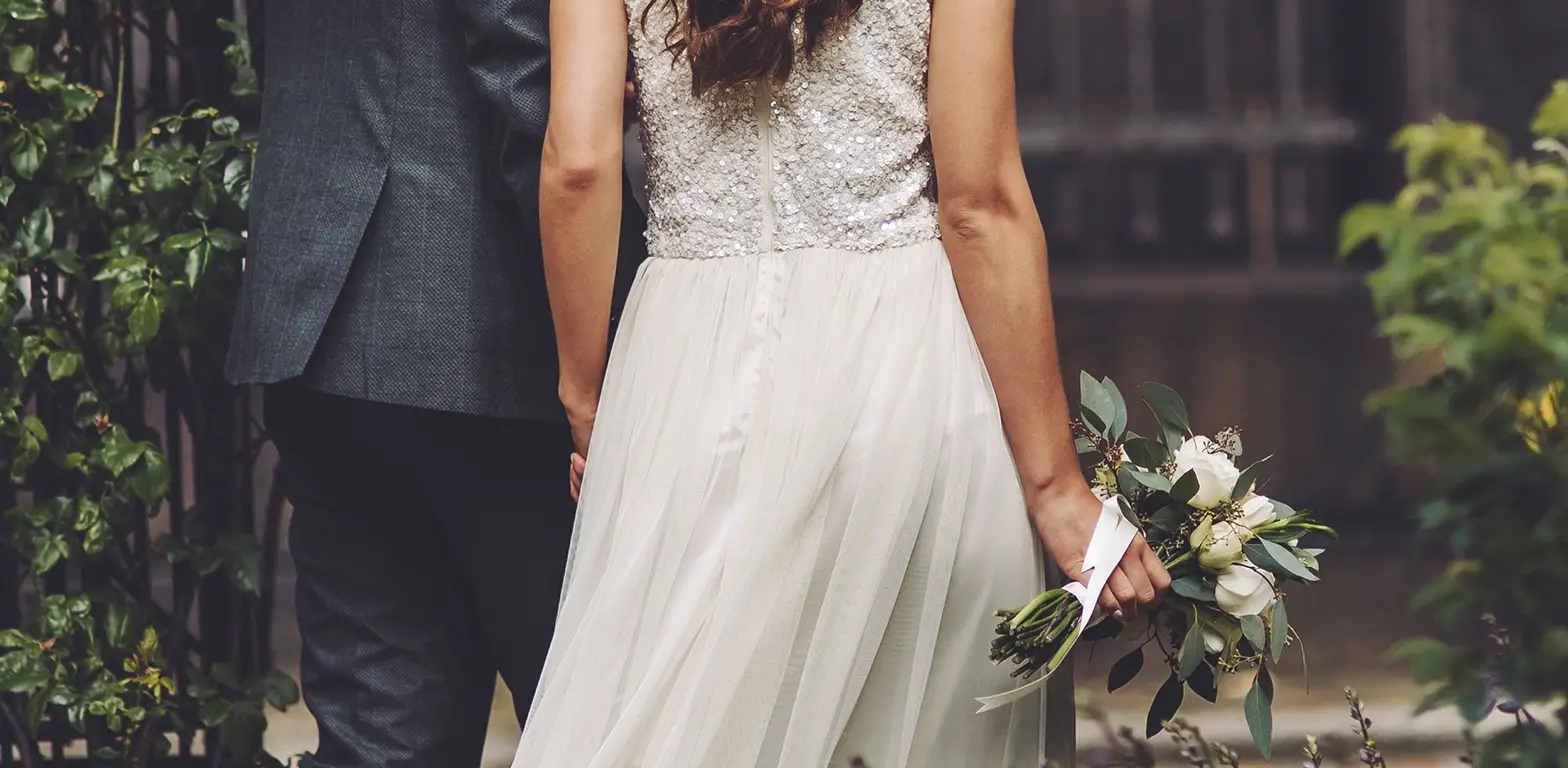 Rückansicht eines Brautpaares. Die Braut im weißen Paillettenkleid hält einen Strauß weißer Rosen, der Bräutigam trägt einen dunklen Anzug. Symbolbild für rechtliche Absicherung und Ehevertrag in Mettmann.